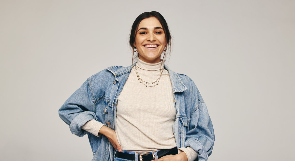 Smiling young woman standing alone in a studio. Happy young woman looking at the camera cheerfully while standing against a grey background. Stylish young woman wearing a denim jacket.