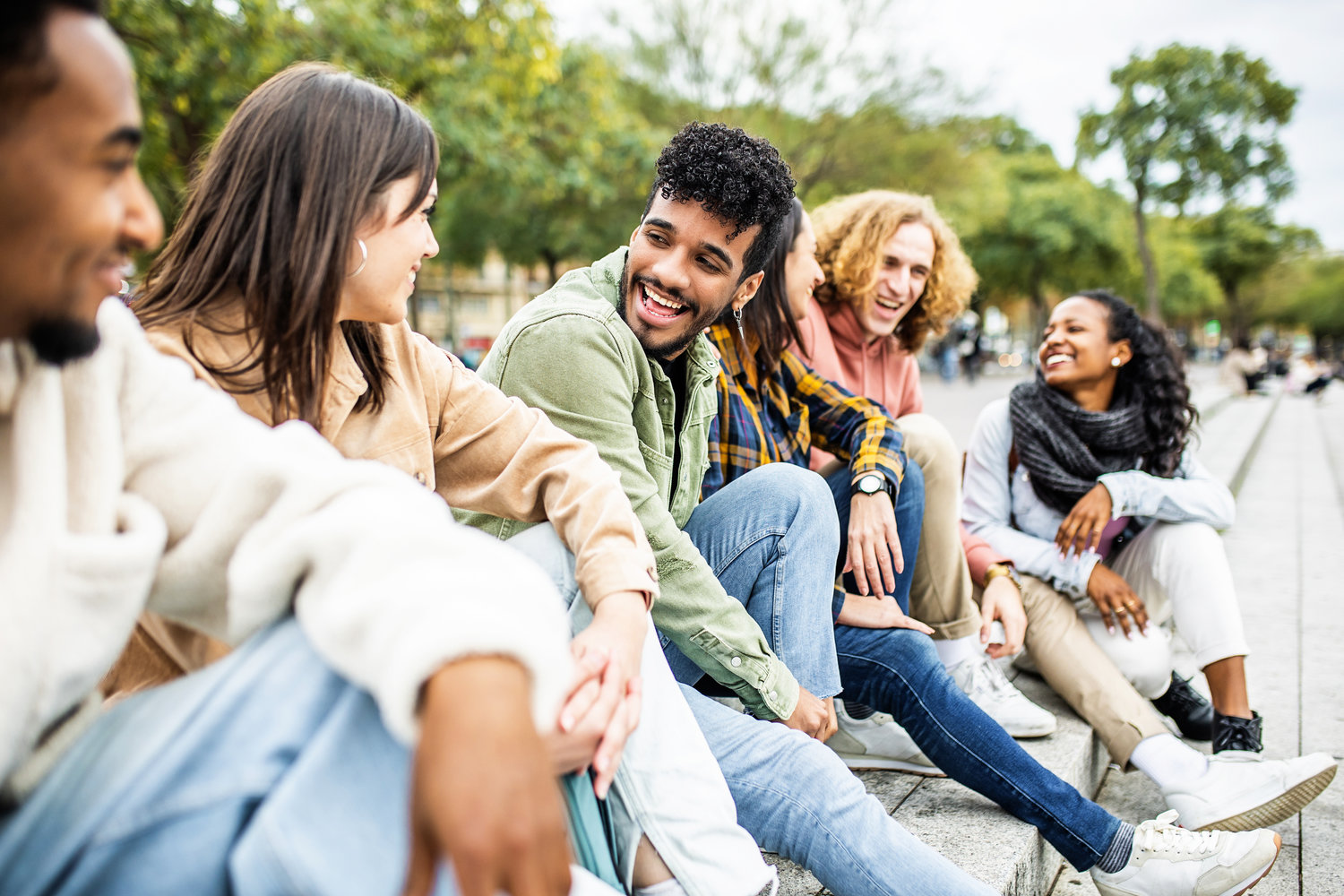 Happy group of trendy young people laughing sitting in city street. Millennial diverse student friends having fun together outdoor