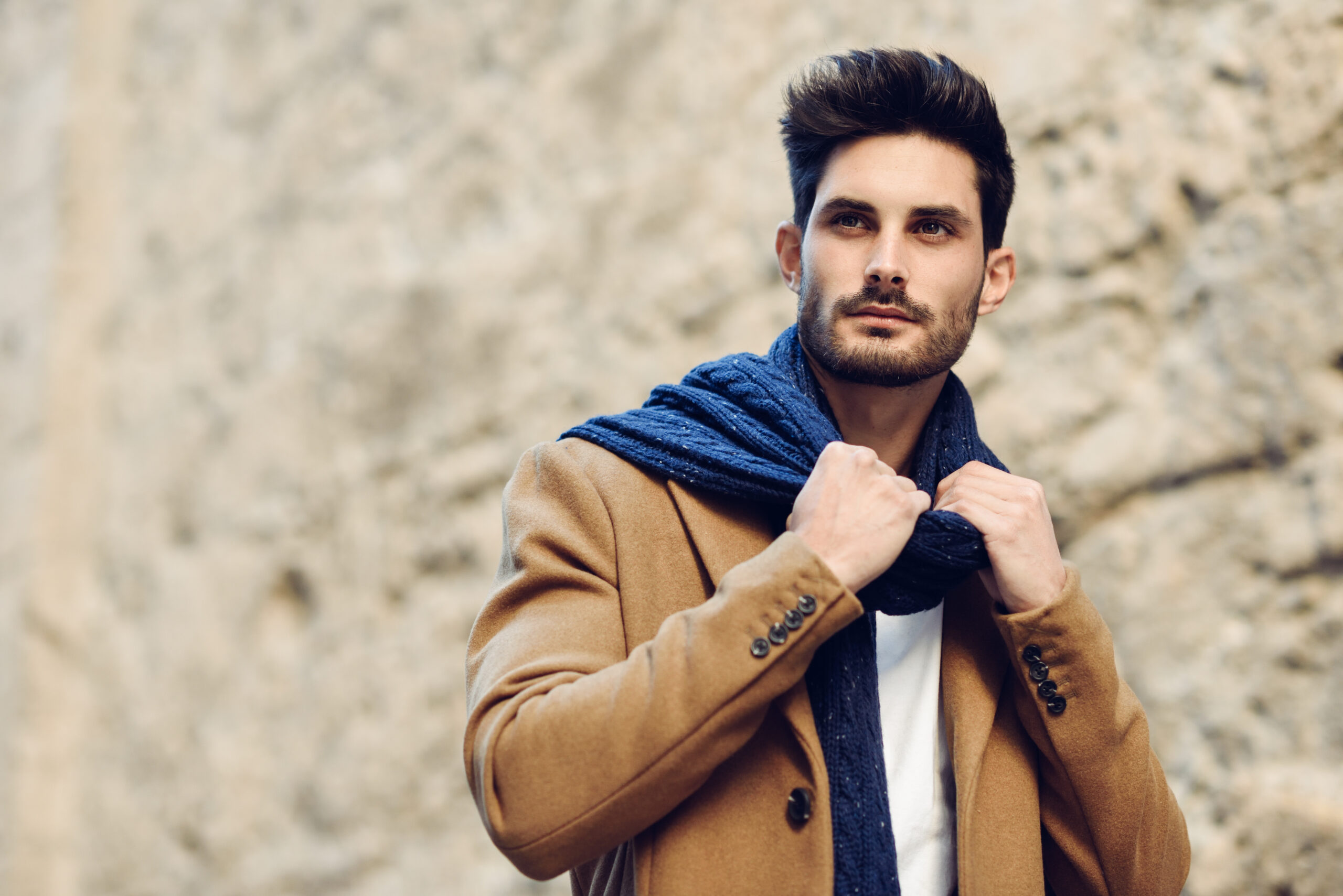 Young man wearing winter clothes in the street. Young bearded guy with modern hairstyle with coat, scarf, blue jeans and t-shirt.