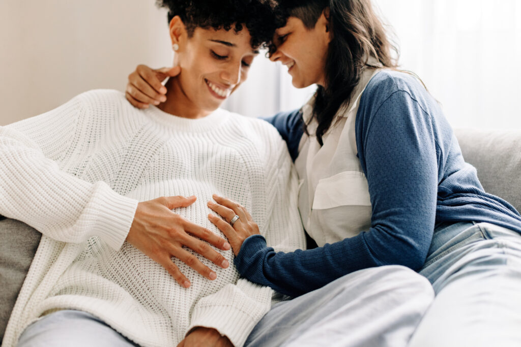 Smiling pregnant couple bonding while sitting together. Happy young woman embracing her pregnant wife in their living room. Pregnant lesbian couple spending quality time together.