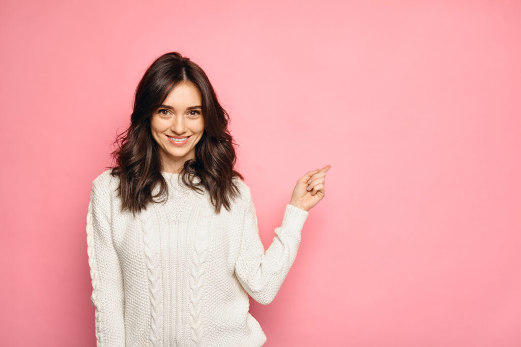 Cute young woman points a finger away on pink colorful background. Girl with brown hair wearing a sweater showing copy space isolated