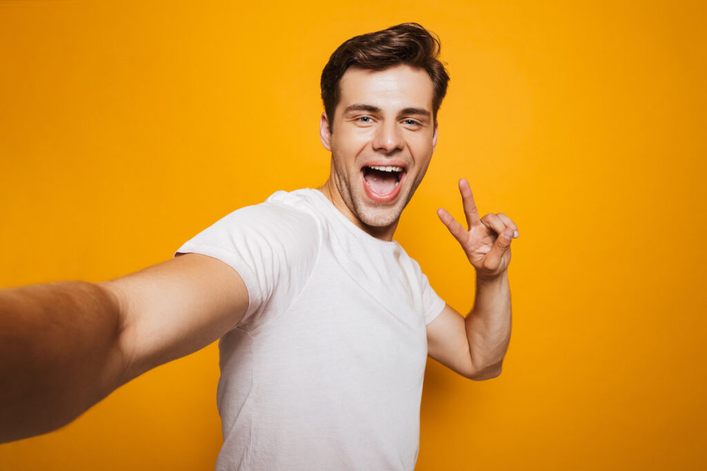 Portrait of a happy young man taking a selfie with outsretched hand, showing peace gesture isolated over yellow background