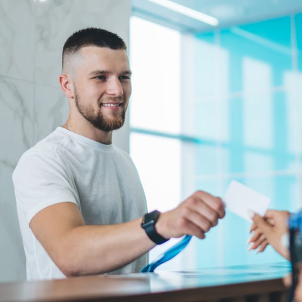 Handsome caucasian man receptionist in gym greeting female client giving key to access for training, happy male employee communicating with 20s woman taking card for opening locker room