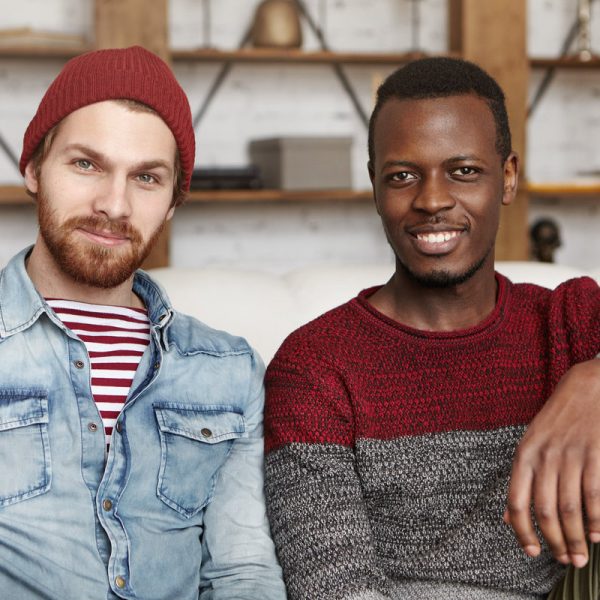 International friendship concept. Indoor shot of two stylish young men having rest in modern cafe interior: handsome bearded hipster in hat and denim jacket sitting next to his African-American friend