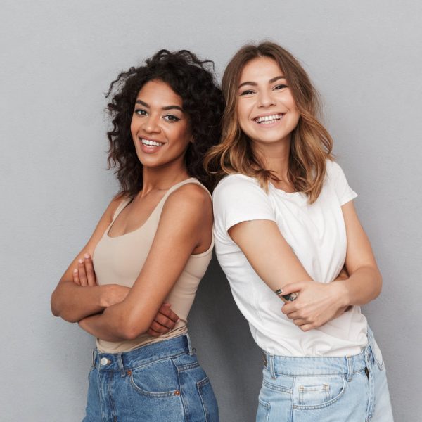 Portrait of two cheerful young women standing together and looking at camera isolated over gray background