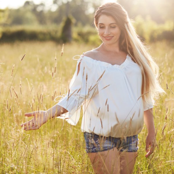 Positive young female anticipates for baby, strolls across wheat field, enjoys sunny day, touches green grass. Outdoor shot of pregnant woman dressed in white blouse and denim shorts. Pregnancy