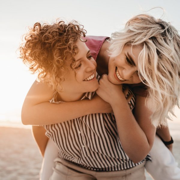 Laughing young woman giving her girlfriend a piggyback while having fun together at a beach at sunset