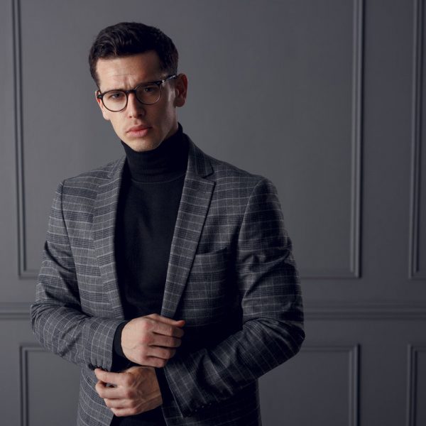 A strong,handsome man in a shawl-gray jacket and black turtleneck, standing in profile with his hands in his pockets and looking up confident.Horizontal view on the gray wall background.