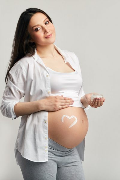 Pregnant woman in third trimester, dressed in comfotable home wear, posing against studio purple wall, looks happily at camera, being in anticipation of baby. Happy pregnancy and motherhood concept