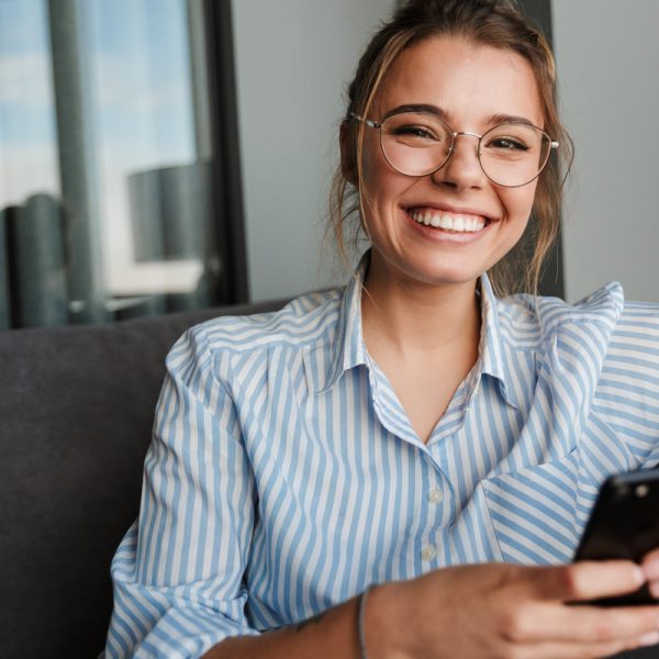 Image of happy young woman in eyeglasses smiling and using cellphone while sitting on couch