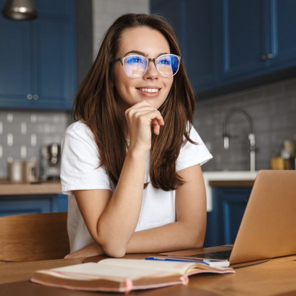 Image of smiling nice woman in eyeglasses working with laptop while sitting at cozy kitchen