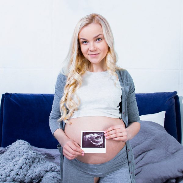 Close up pregnant beautiful woman waiting for a child holding ultrasound baby picture close to beautiful belly, tummy. Laying on gray bed sheets. Preparation for childbirth maternity parents concept