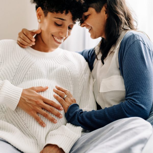 Smiling pregnant couple bonding while sitting together. Happy young woman embracing her pregnant wife in their living room. Pregnant lesbian couple spending quality time together.