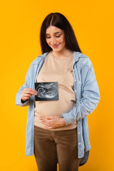 Excited young pregnant woman embracing belly and showing ultrasound picture of unborn baby on yellow backgorund. Exam, motherhood, family, love, expecting baby